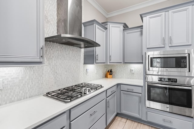 kitchen featuring gray cabinets, backsplash, stainless steel appliances, ornamental molding, and wall chimney exhaust hood