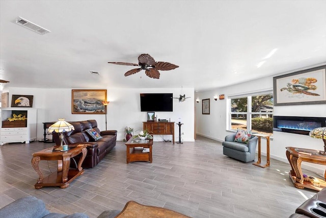 living room with a ceiling fan, wood tiled floor, visible vents, and a glass covered fireplace