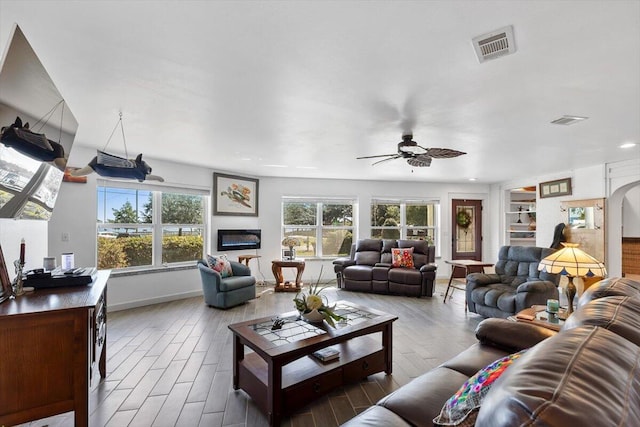 living area featuring wood finished floors, a glass covered fireplace, visible vents, and a ceiling fan