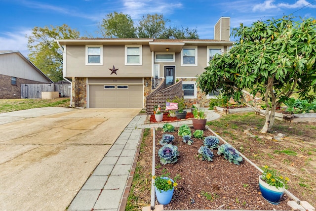 bi-level home with an attached garage, fence, concrete driveway, a vegetable garden, and a chimney