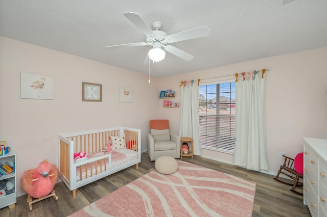 bedroom featuring ceiling fan, a nursery area, dark wood-type flooring, and a textured ceiling