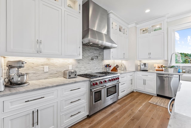 kitchen featuring white cabinetry, light wood-type flooring, stainless steel appliances, light stone countertops, and wall chimney range hood