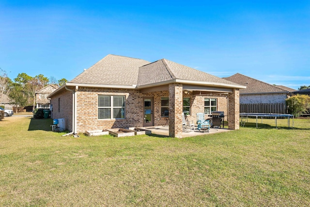 back of property featuring a trampoline, brick siding, a patio, roof with shingles, and a lawn