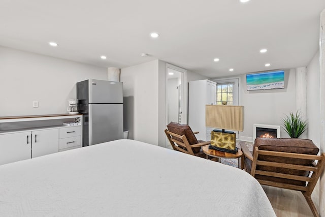 bedroom featuring a lit fireplace, light wood-style flooring, freestanding refrigerator, and recessed lighting