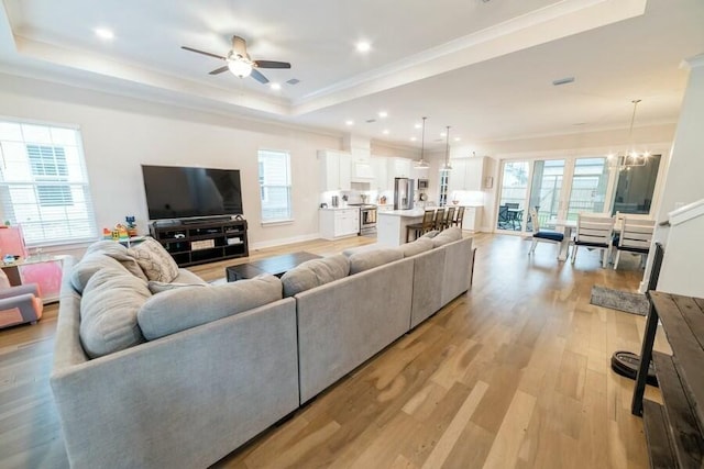 living area featuring light wood finished floors, ceiling fan with notable chandelier, a tray ceiling, and ornamental molding