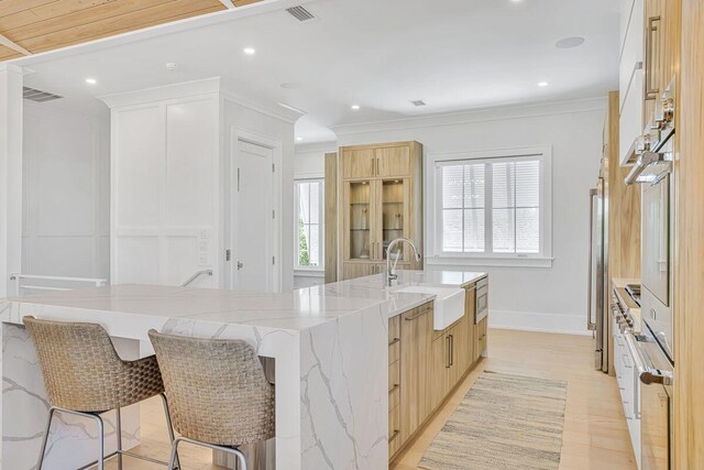 kitchen with a sink, light stone counters, light brown cabinetry, ornamental molding, and a breakfast bar