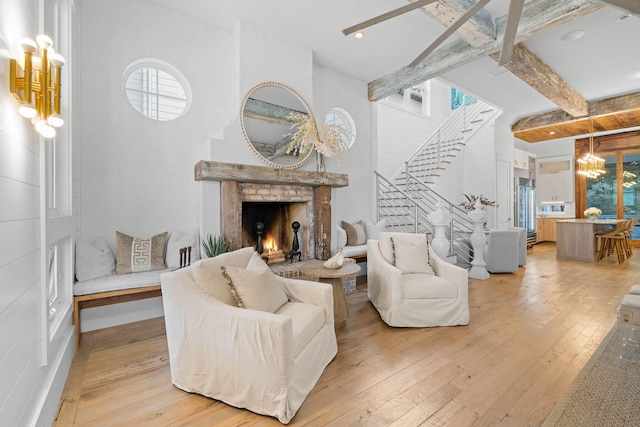 living area featuring a warm lit fireplace, beamed ceiling, stairs, light wood-style floors, and a chandelier