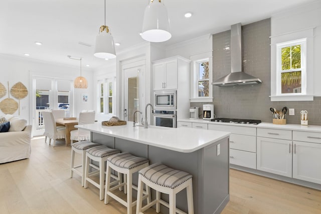 kitchen featuring a kitchen island with sink, appliances with stainless steel finishes, backsplash, wall chimney exhaust hood, and crown molding