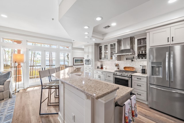 kitchen featuring a kitchen island, appliances with stainless steel finishes, wall chimney exhaust hood, a kitchen bar, and glass insert cabinets