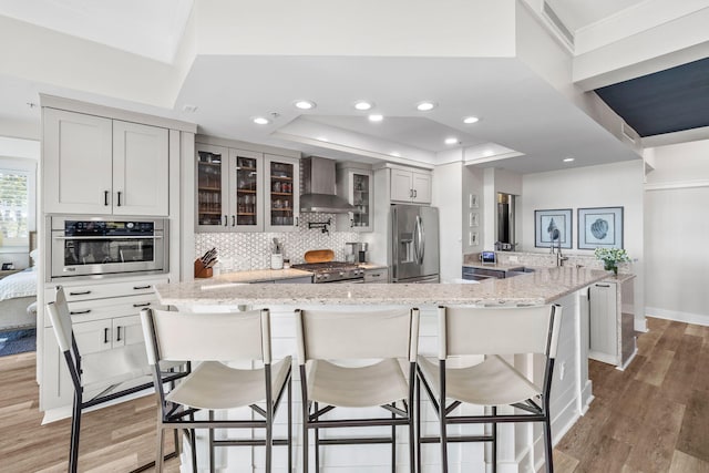 kitchen with light stone counters, a breakfast bar area, stainless steel appliances, glass insert cabinets, and wall chimney range hood