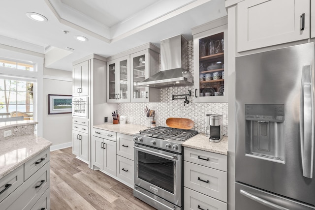 kitchen featuring stainless steel appliances, light stone countertops, wall chimney exhaust hood, tasteful backsplash, and glass insert cabinets