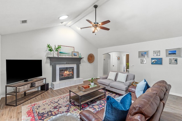 living room featuring arched walkways, vaulted ceiling with beams, a tile fireplace, wood finished floors, and baseboards