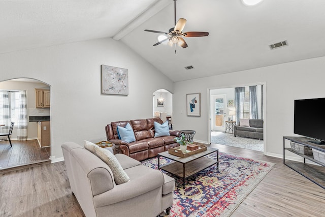 living area featuring arched walkways, visible vents, a ceiling fan, light wood finished floors, and beamed ceiling