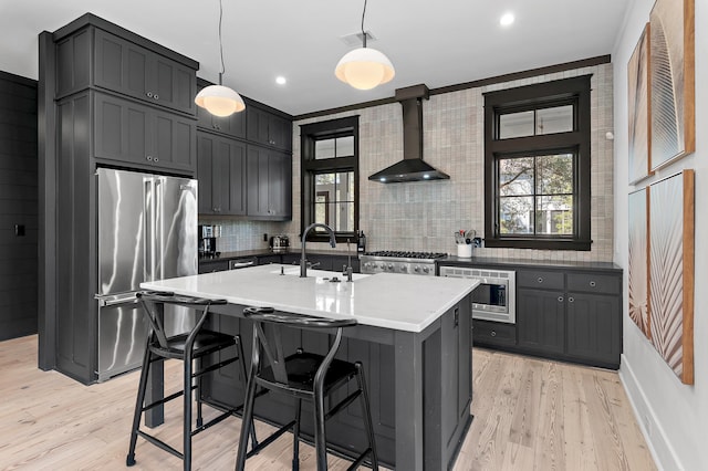 kitchen with stainless steel appliances, a sink, wall chimney exhaust hood, an island with sink, and pendant lighting