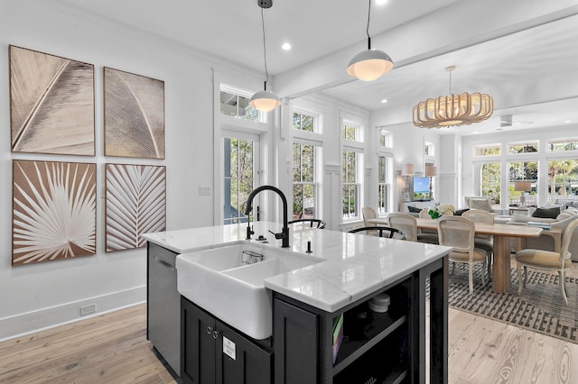 kitchen with decorative light fixtures, a kitchen island with sink, a sink, light stone countertops, and dark cabinets