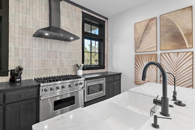 kitchen featuring stainless steel appliances, backsplash, a sink, wall chimney range hood, and dark cabinets