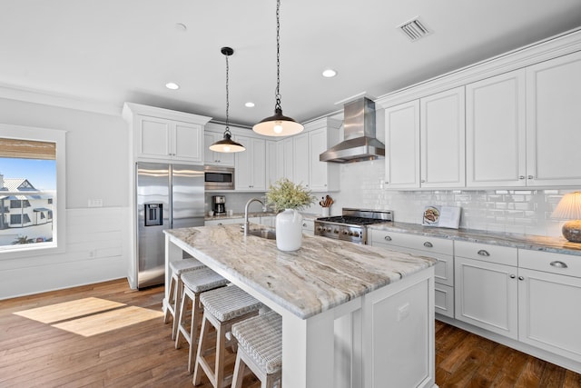 kitchen with appliances with stainless steel finishes, white cabinets, a center island with sink, and wall chimney exhaust hood