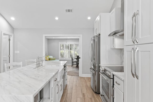 kitchen featuring wall chimney range hood, white cabinets, light wood-type flooring, and appliances with stainless steel finishes