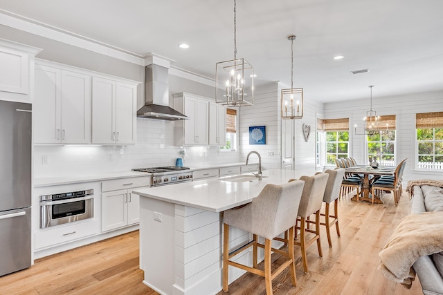 kitchen with a breakfast bar area, stainless steel appliances, light wood-style flooring, a sink, and wall chimney range hood