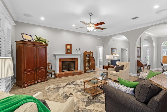 living area featuring light wood-type flooring, visible vents, arched walkways, crown molding, and a brick fireplace