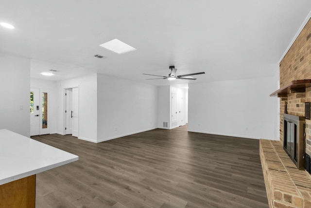 unfurnished living room featuring ceiling fan, a skylight, visible vents, a brick fireplace, and dark wood finished floors