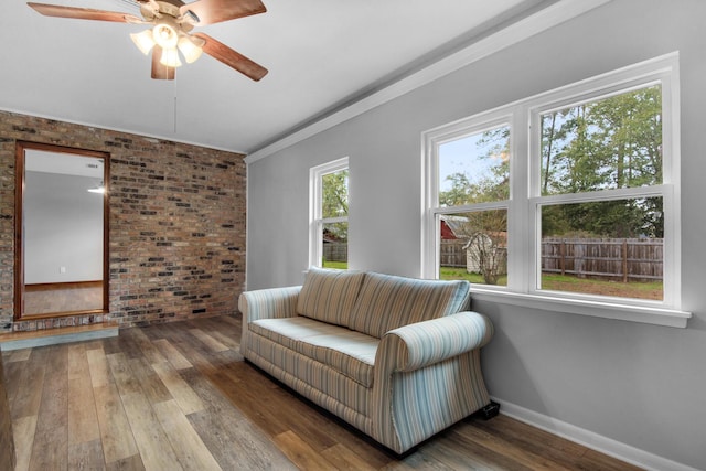 living area with ornamental molding, a ceiling fan, hardwood / wood-style flooring, brick wall, and baseboards