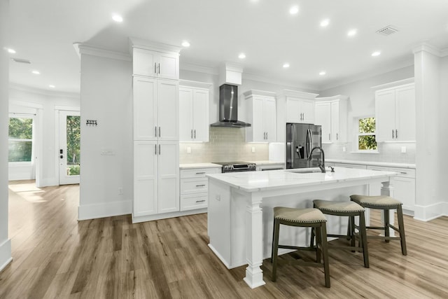 kitchen featuring crown molding, appliances with stainless steel finishes, a kitchen island with sink, a sink, and wall chimney exhaust hood