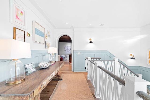 hallway with arched walkways, a wainscoted wall, crown molding, a decorative wall, and light wood-style flooring