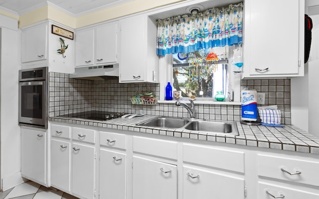 kitchen featuring tile countertops, ornamental molding, oven, under cabinet range hood, and a sink