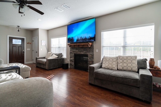 living area with a textured ceiling, a stone fireplace, wood finished floors, visible vents, and a ceiling fan