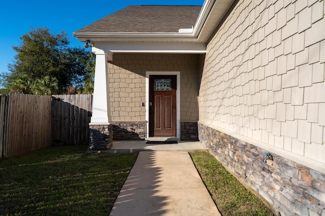 view of exterior entry with stone siding, roof with shingles, fence, and a lawn