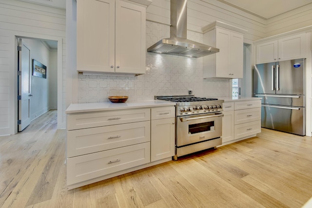 kitchen with wall chimney exhaust hood, appliances with stainless steel finishes, light countertops, light wood-type flooring, and white cabinetry