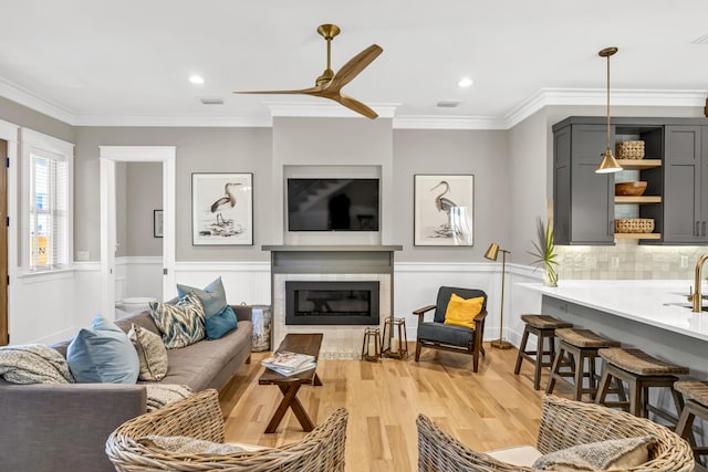 living area featuring ornamental molding, a glass covered fireplace, wainscoting, and light wood-style flooring