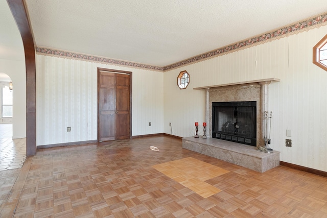 unfurnished living room featuring wallpapered walls, baseboards, a fireplace, arched walkways, and a textured ceiling