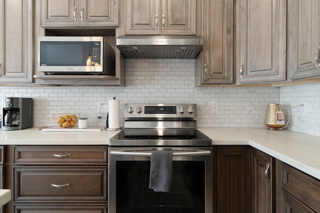 kitchen featuring light stone countertops, dark brown cabinetry, under cabinet range hood, appliances with stainless steel finishes, and backsplash