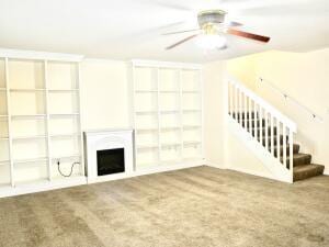 unfurnished living room with built in features, carpet, a fireplace, a ceiling fan, and stairs