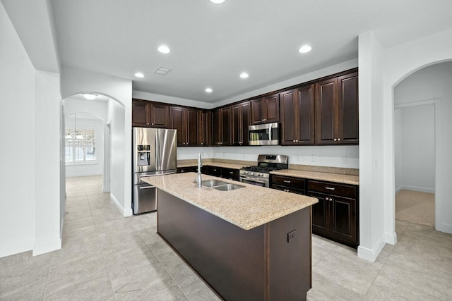 kitchen with dark brown cabinetry, a center island with sink, arched walkways, stainless steel appliances, and a sink