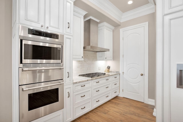 kitchen featuring light wood finished floors, ornamental molding, stainless steel appliances, white cabinetry, and wall chimney range hood