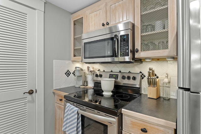 kitchen with tasteful backsplash, dark countertops, appliances with stainless steel finishes, glass insert cabinets, and light brown cabinetry