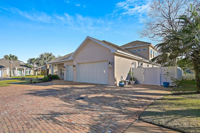 view of side of property with an attached garage, brick siding, fence, decorative driveway, and a gate