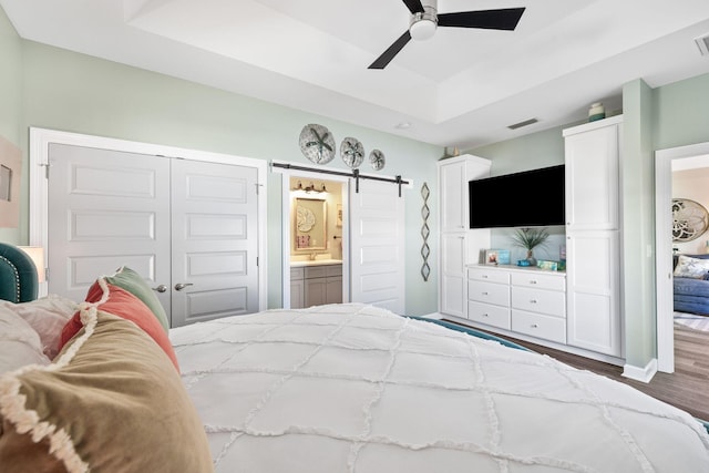 bedroom featuring a barn door, a raised ceiling, connected bathroom, a ceiling fan, and wood finished floors