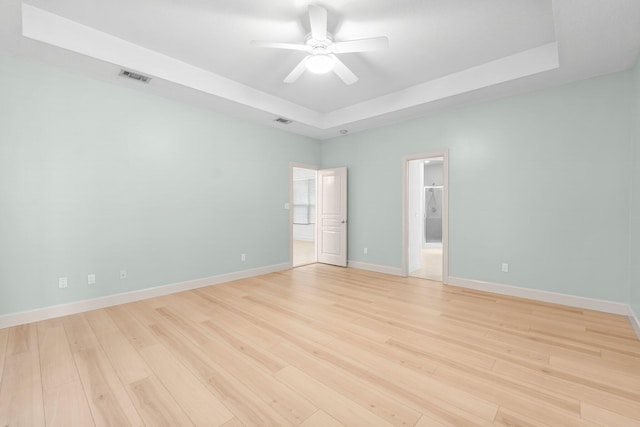 empty room featuring a tray ceiling, light wood finished floors, visible vents, ceiling fan, and baseboards