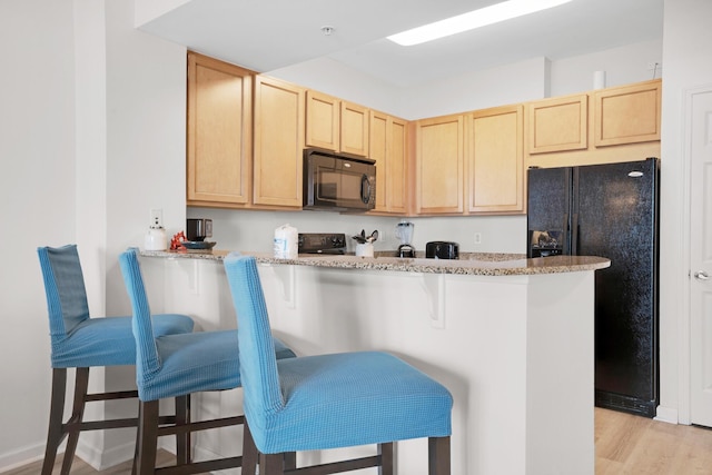 kitchen featuring light wood-style flooring, a breakfast bar, light stone countertops, black appliances, and light brown cabinets