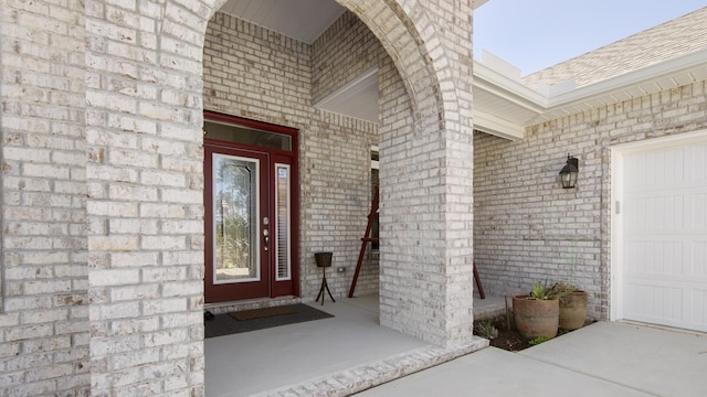 view of exterior entry featuring an attached garage, a shingled roof, and brick siding