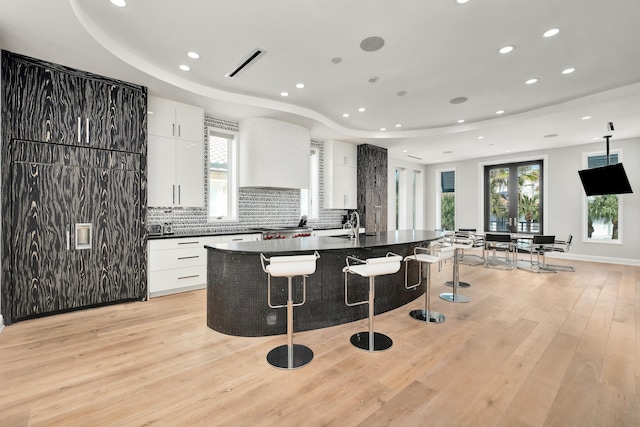 kitchen with light wood-style flooring, a kitchen breakfast bar, backsplash, dark countertops, and a center island with sink