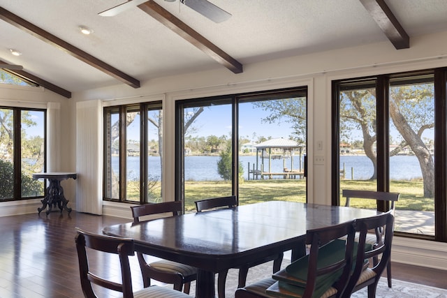 dining space with a water view, dark wood-type flooring, a ceiling fan, lofted ceiling with beams, and a textured ceiling