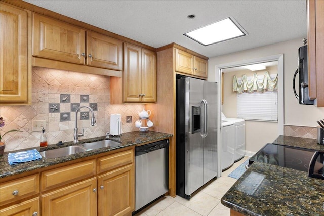 kitchen featuring washing machine and clothes dryer, appliances with stainless steel finishes, light tile patterned flooring, a sink, and dark stone countertops