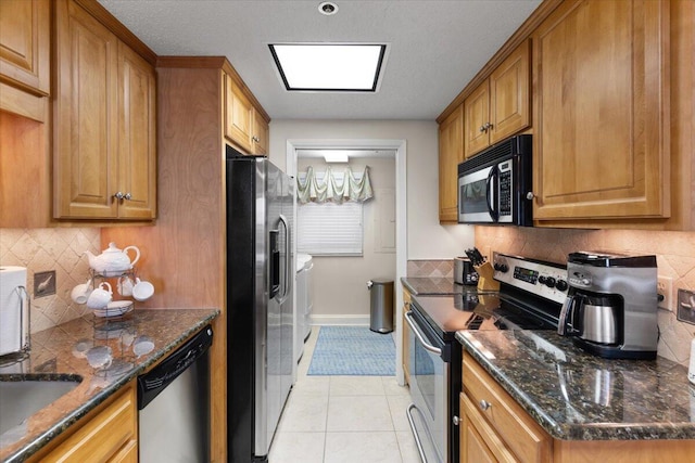 kitchen with brown cabinets, washer and clothes dryer, stainless steel appliances, light tile patterned flooring, and dark stone countertops