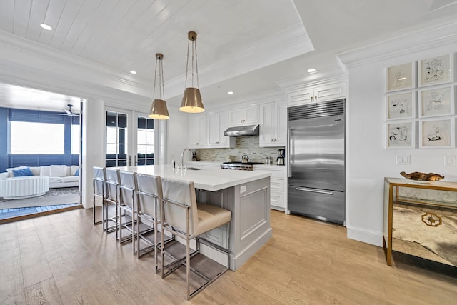 kitchen with a tray ceiling, crown molding, under cabinet range hood, a kitchen bar, and built in fridge