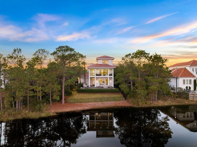 back of property at dusk with a fenced front yard, a water view, a lawn, and a balcony